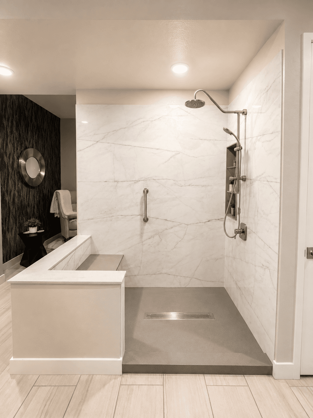 Modern bathroom with walk-in shower featuring marble tile walls, rainfall showerhead, and handheld sprayer; brushed nickel fixtures; gray floor pan with linear drain; white vanity visible on left; black accent wall with round mirror in background